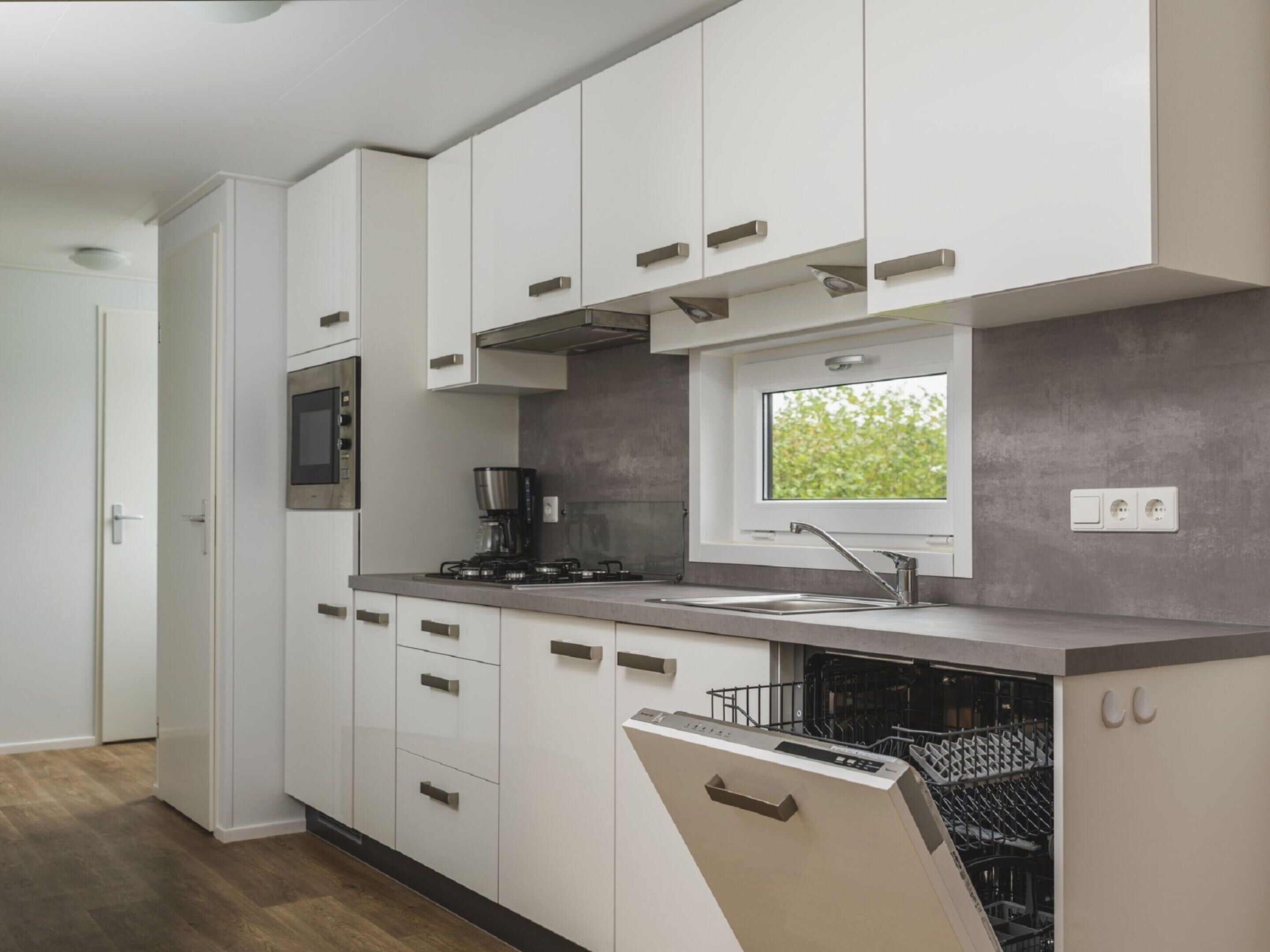 A modern kitchen with white cabinets, a built-in oven, a coffee maker, a window, and a dishwasher.
