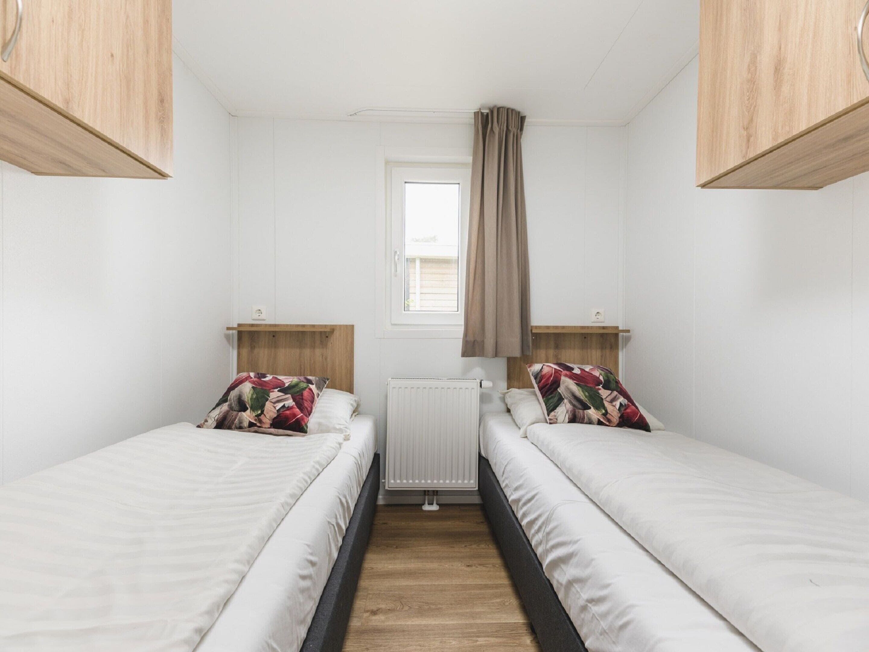 A small, clean room with two single beds, wooden headboards, a window with a curtain, and a radiator.