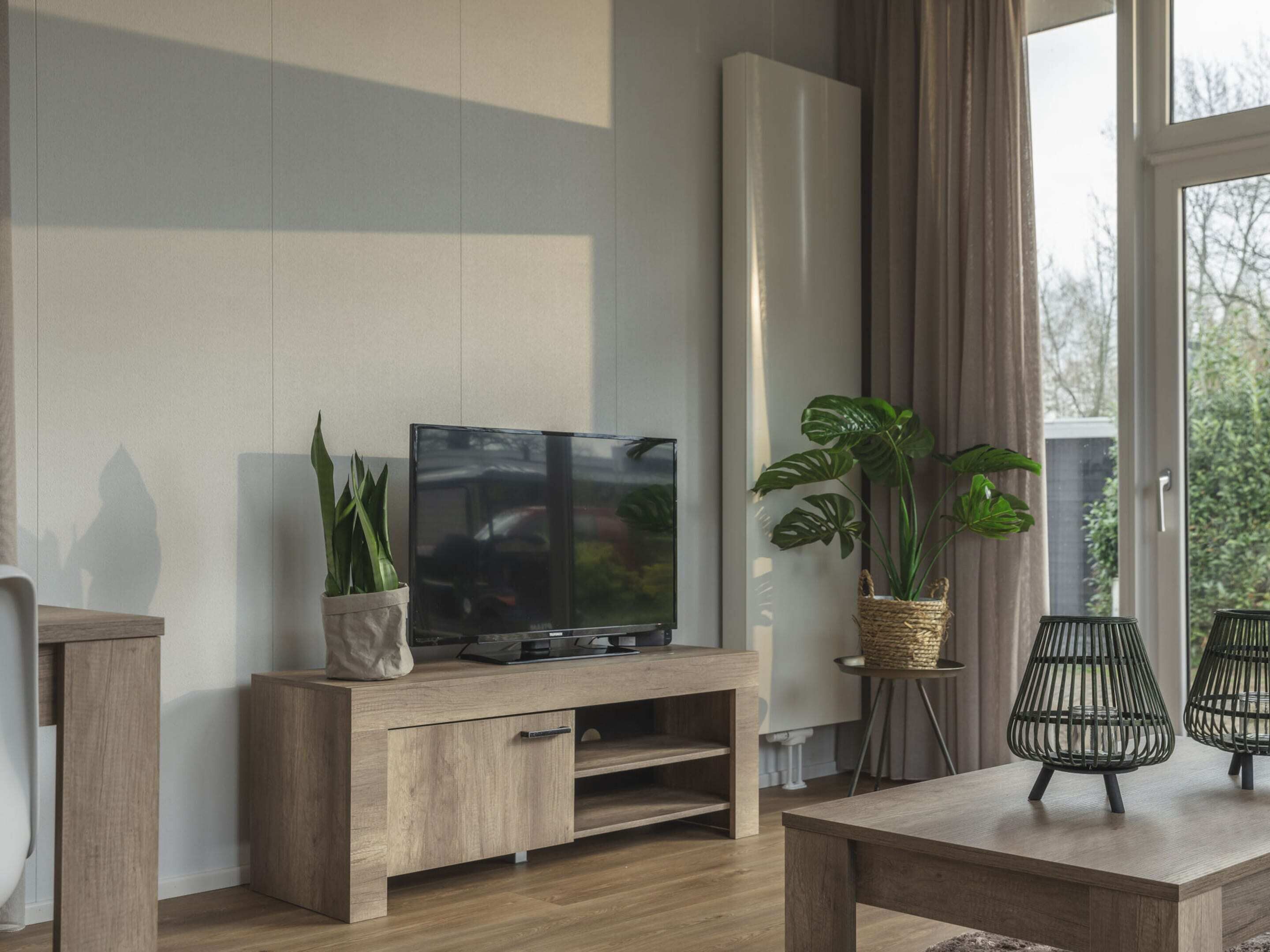 Un salon moderne avec un meuble TV en bois, un téléviseur à écran plat, deux plantes en pot et une petite table avec deux chaises.