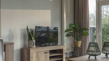Un salon moderne avec un meuble TV en bois, un téléviseur à écran plat, deux plantes en pot et une petite table avec deux chaises.