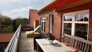A wooden deck with a table and chairs, a red roof, and a brick building with large windows. - Penthuus Modern Retreat (Juist)