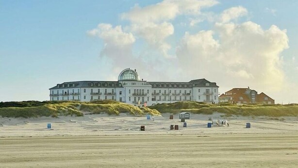 A large, multi-story building with a prominent dome structure, situated on a sandy beach with dunes in the background. - Penthuus Modern Retreat (Juist)