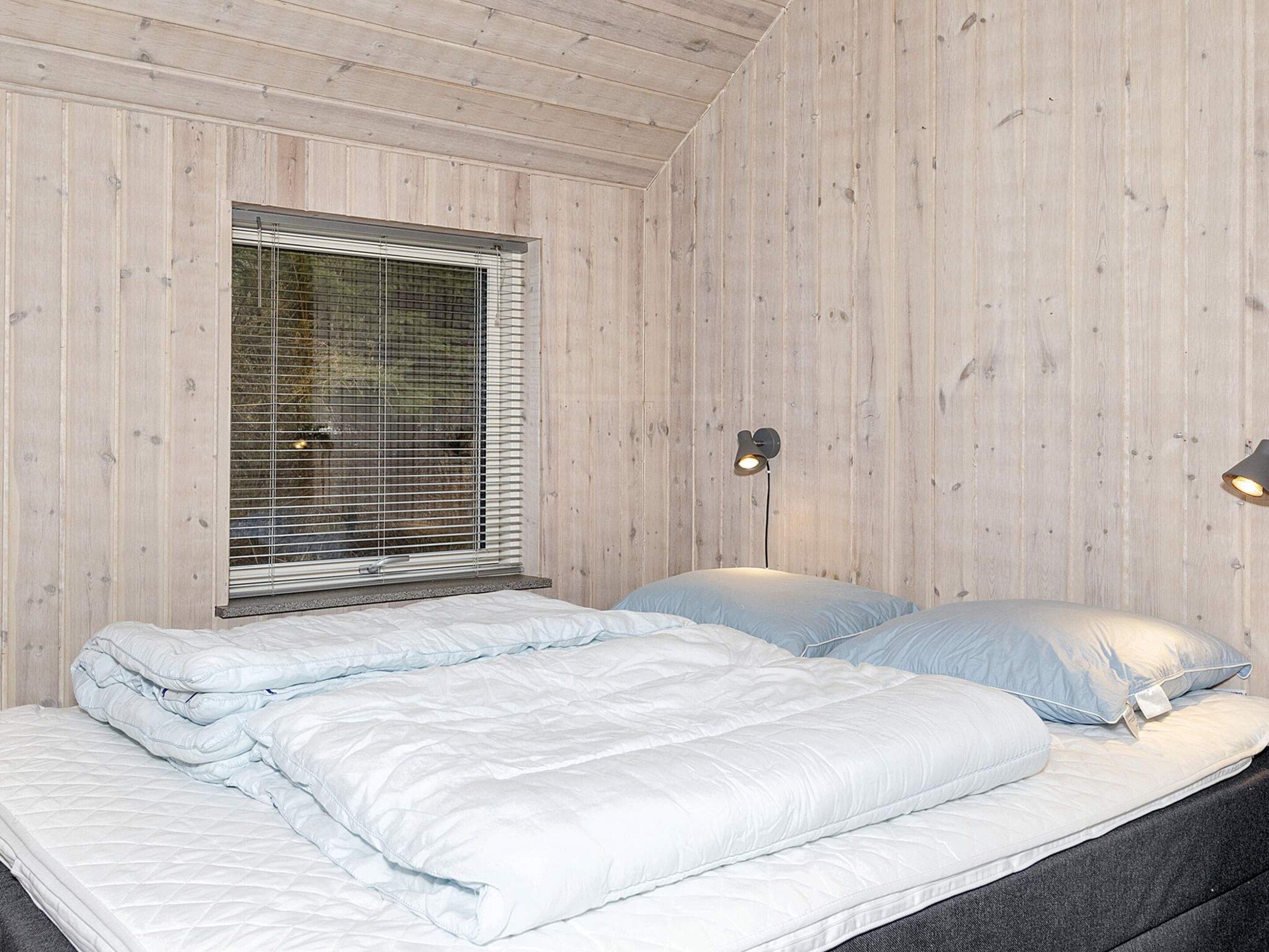 A bedroom with two beds, wooden walls, a window with blinds, and two wall-mounted lamps.