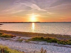 En strand med en solnedgång över vattnet, en sandstrand och gräsklädda sanddyner.