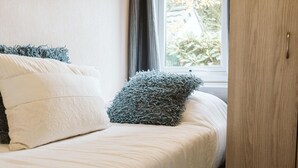 Una cama con ropa de cama blanca y dos almohadas, una blanca y otra gris, en una habitación con ventana y puerta de madera.
