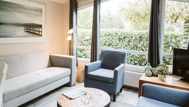 Una sala de estar moderna con sofá, sillón y mesa de centro. Hay un cuadro en la pared, una lámpara y un televisor sobre una mesa de madera.