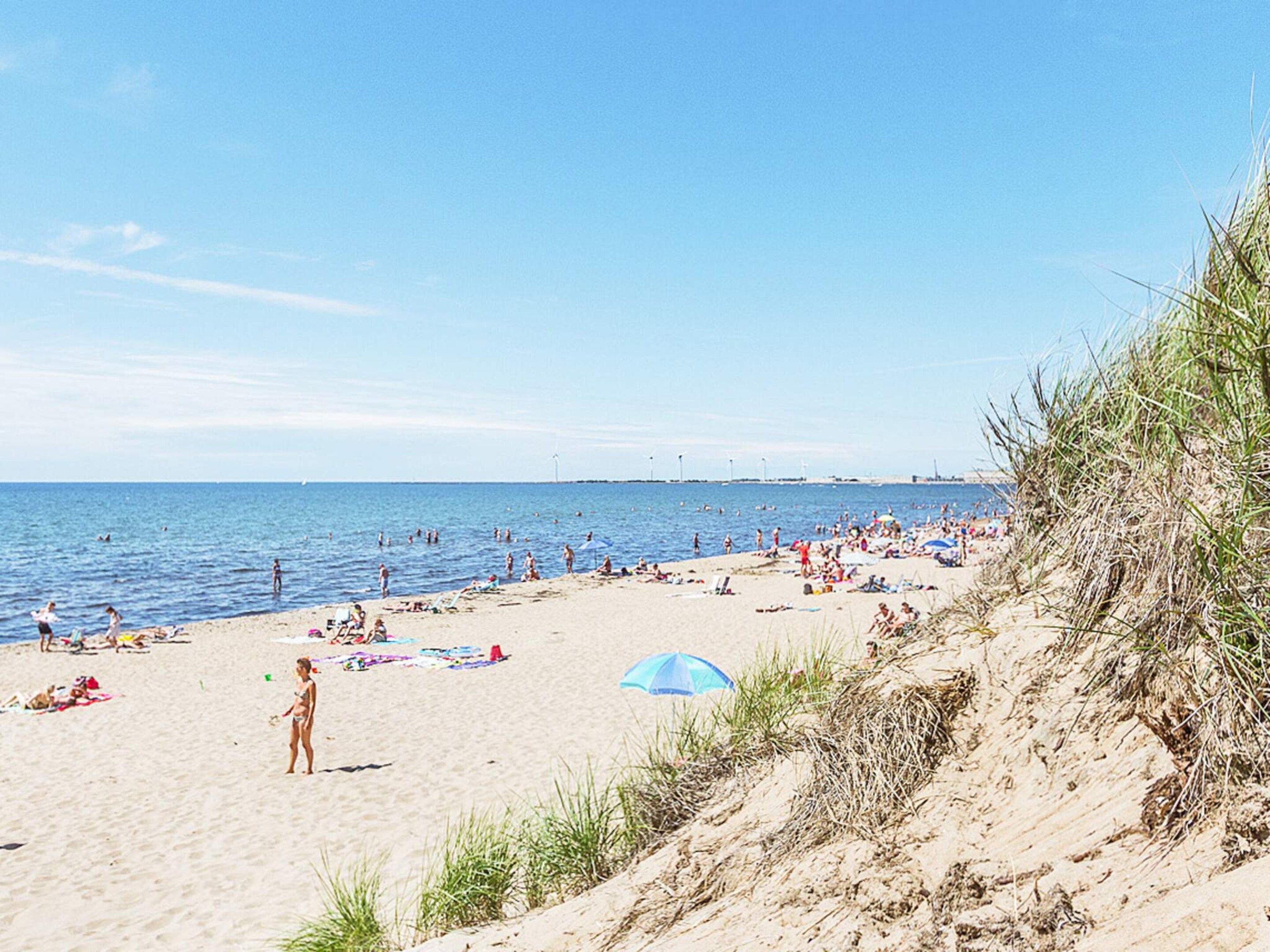 A beach with people sunbathing and swimming, a clear sky, and a distant view of a city skyline.