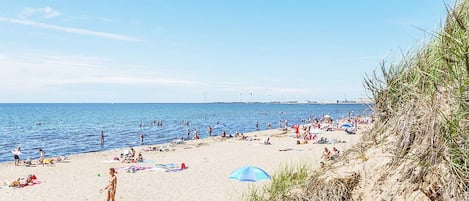 Une plage où des gens se prélassent au soleil et se baignent, un ciel dégagé et une vue lointaine sur les gratte-ciel d’une ville.