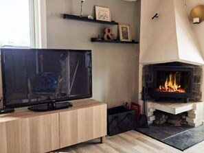 Uma sala de estar com televisão sobre um móvel de madeira, lareira acesa e uma janela.