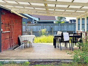 Relax on the covered patio with a table and chairs, surrounded by lush greenery and a charming brick building in the background.
