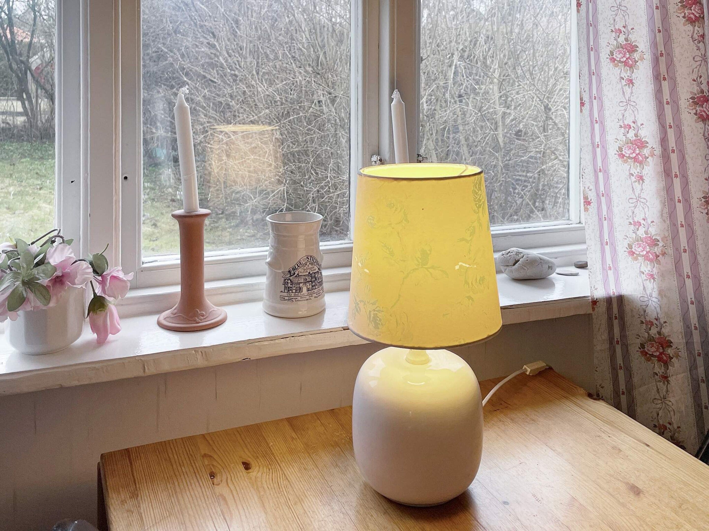 Una habitación con ventana, una mesa, una lámpara, una vela y un florero con flores.