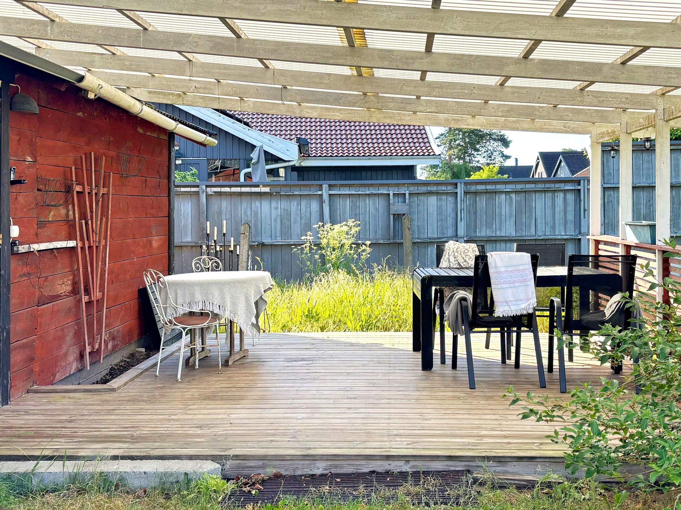 A covered outdoor patio with a table and chairs, a wooden deck, and a red building in the background.