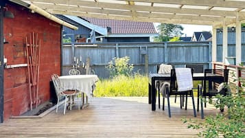 A covered outdoor patio with a table and chairs, a wooden deck, and a red building in the background.