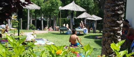 Un jardin agrémenté de palmiers et de parasols en chaume, un espace piscine avec des chaises longues, et des gens qui se détendent.