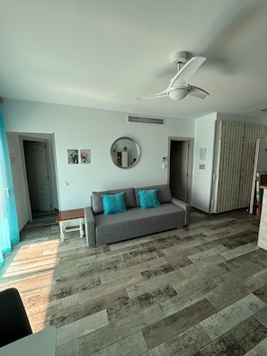 Ein Wohnzimmer mit grauem Sofa, Deckenventilator, rundem Spiegel und Fliesenboden.
