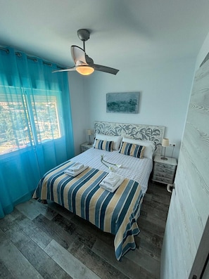 Une chambre à coucher avec un lit, un ventilateur de plafond, une table de chevet, une lampe et une fenêtre avec des rideaux.