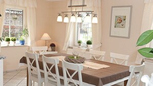 A dining area with a table set for four, surrounded by white chairs, a hanging light fixture, and potted plants on the windowsill. - Casa Schalkenmehren Grande (Schalkenmehren)