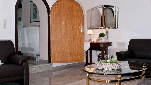 A modern living room with a glass coffee table, two dark sofas, a wooden door, and a small table with a lamp and decorative items. - Casa Schalkenmehren Grande (Schalkenmehren)