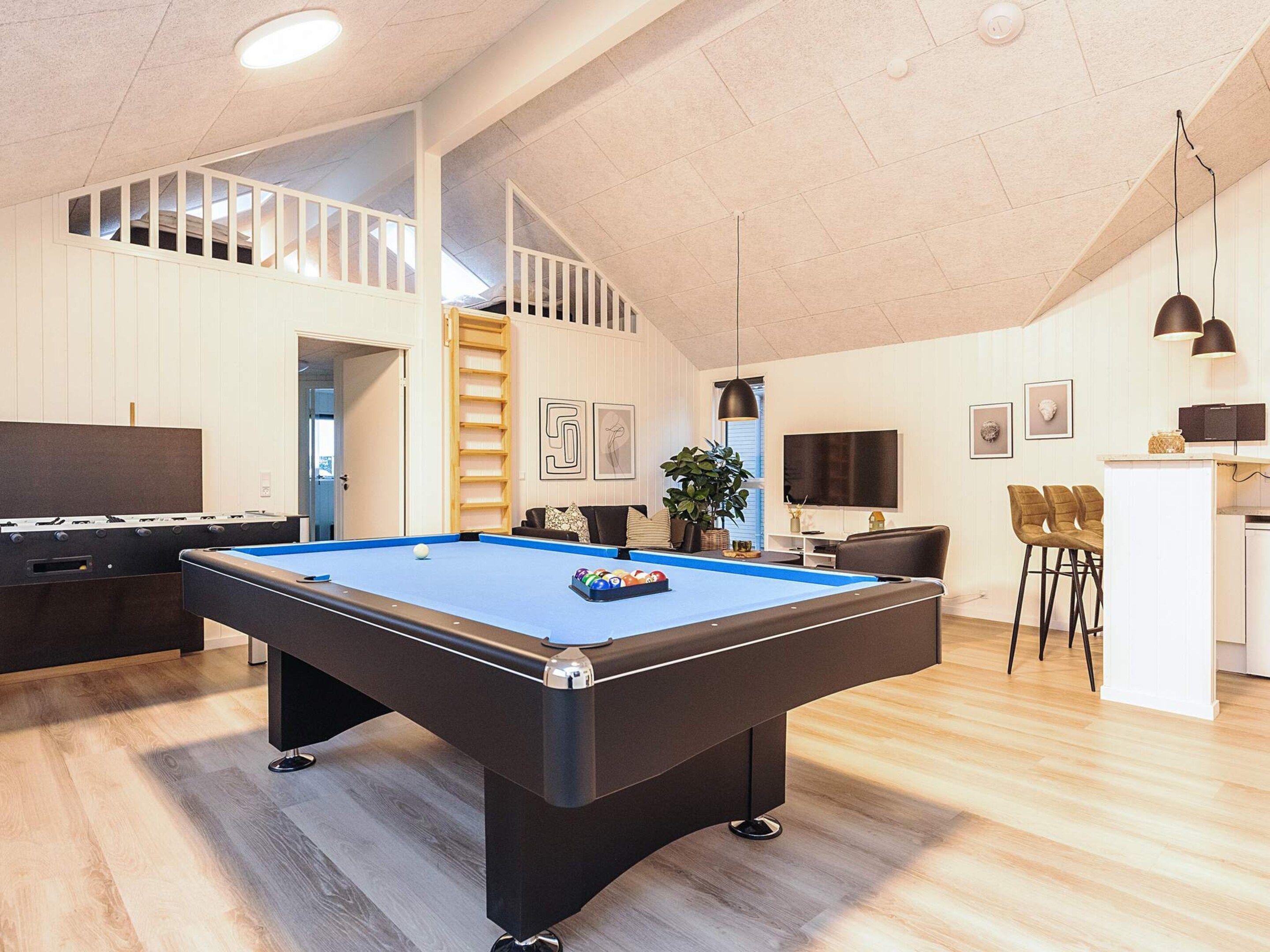 Ein geräumiger Raum mit Billardtisch, Kicker, einer Treppe, einer Küchenzeile und einer Sitzecke mit Stühlen.