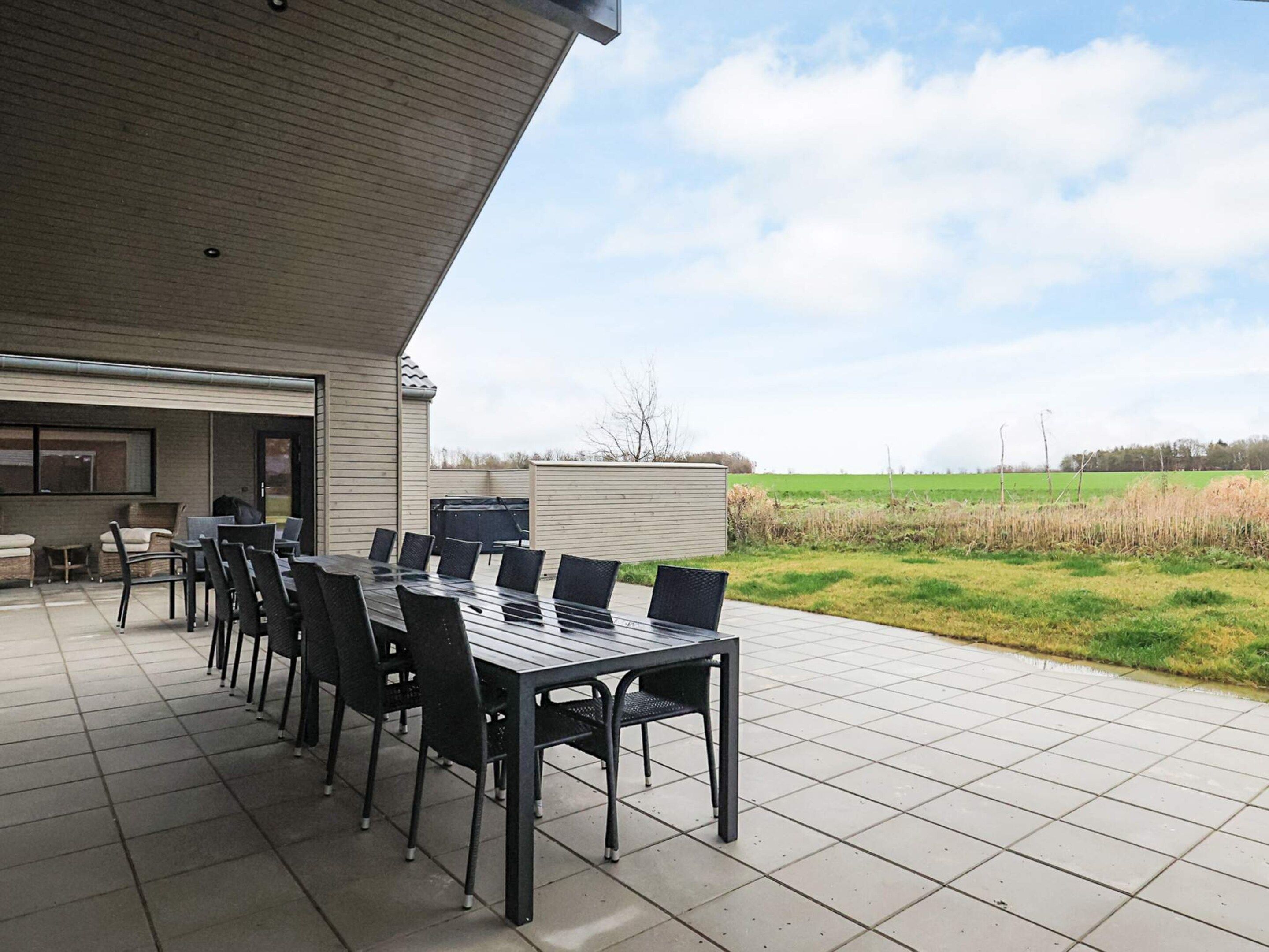 Une terrasse extérieure avec une longue table à manger et des chaises, un barbecue et une vue sur un champ verdoyant.