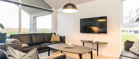 A modern living room with a leather sofa, a coffee table, a flat-screen TV mounted on the wall, and large windows offering a view of the outdoors.