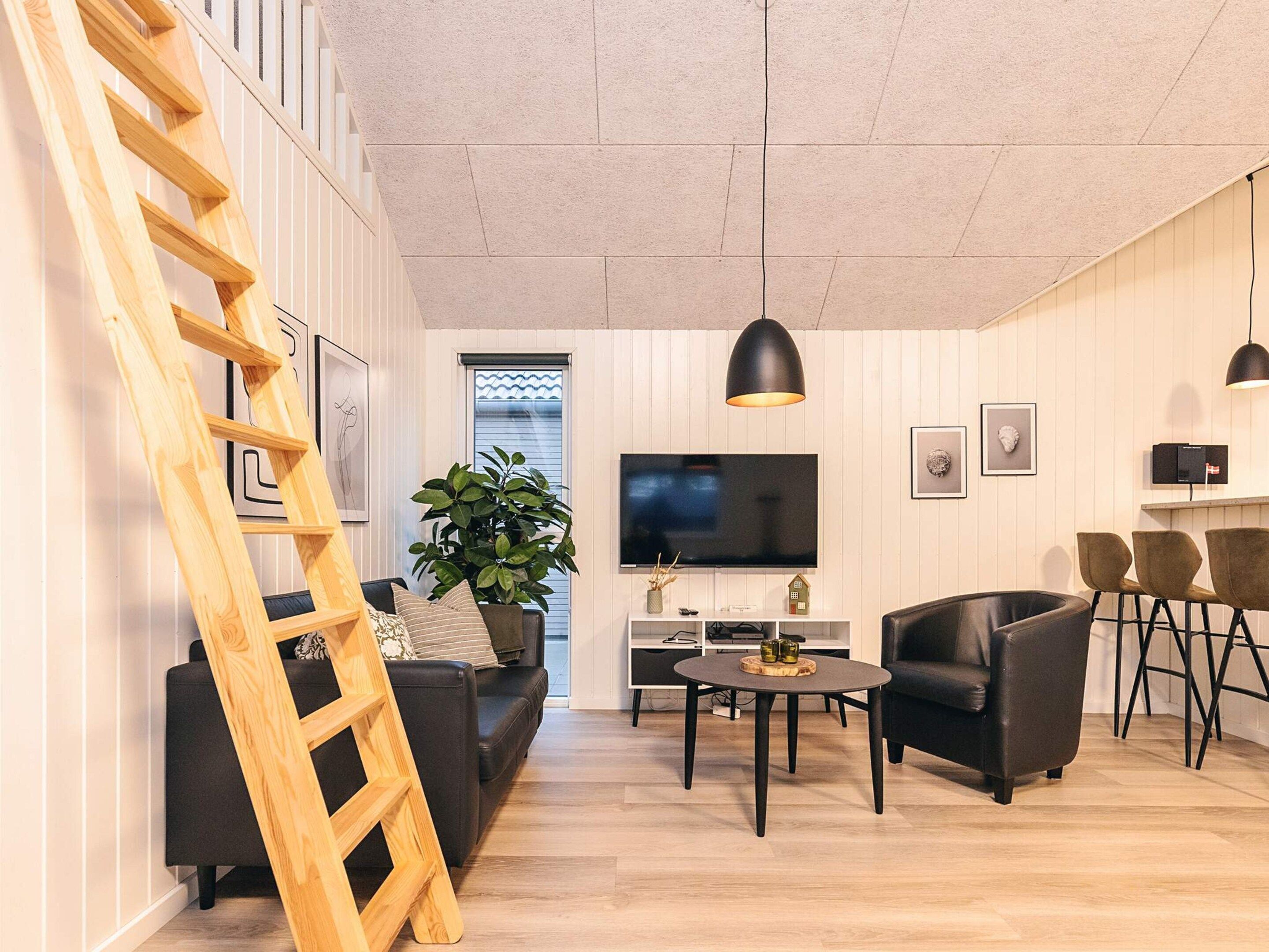 A modern living room with a wooden ladder, a black sofa, a round coffee table, a black leather chair, and a TV mounted on the wall.