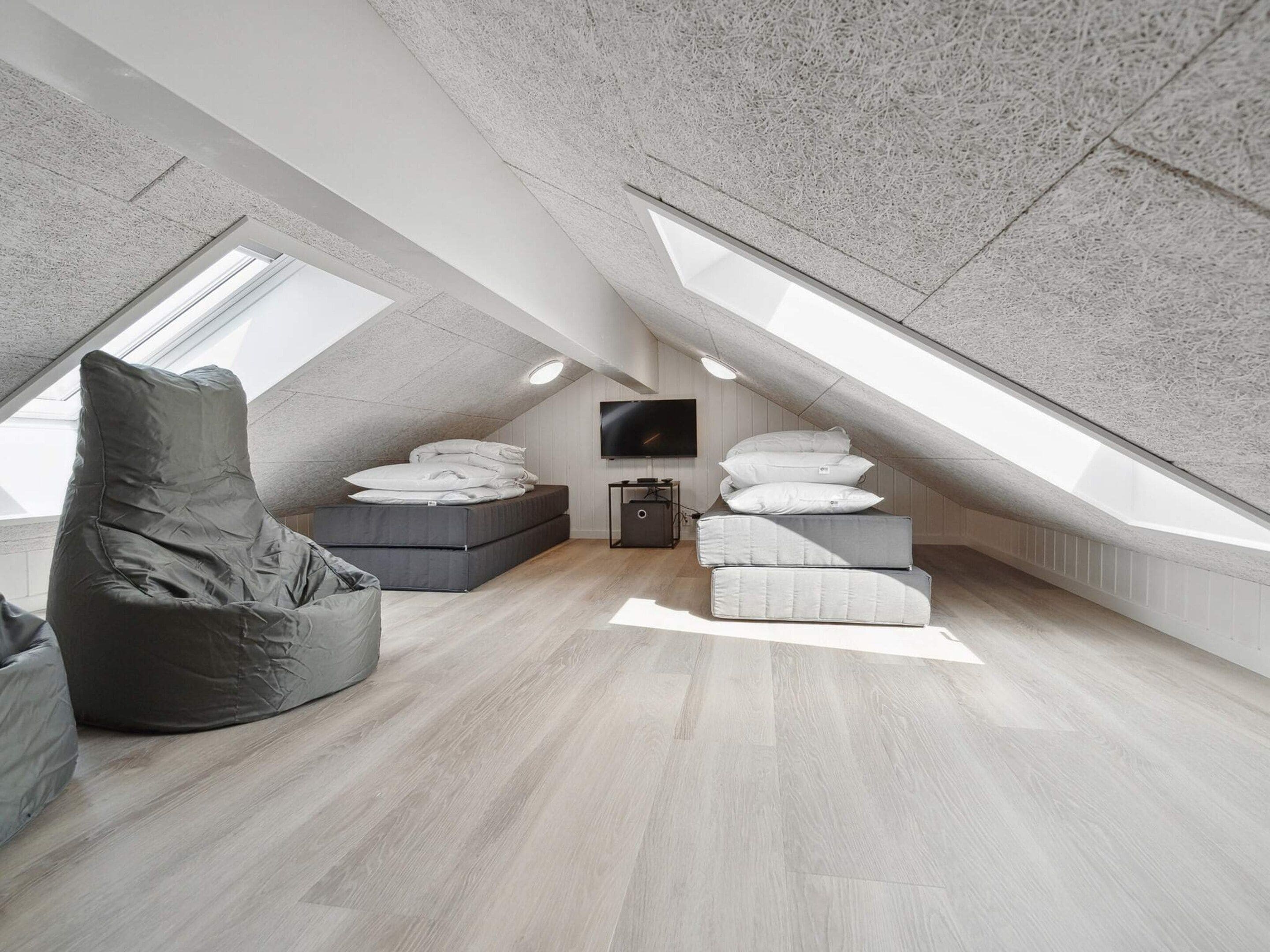 A modern loft bedroom with a sloped ceiling, a large window, a bed with pillows, a bean bag chair, and a television.