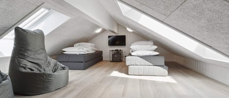 A modern loft bedroom with a sloped ceiling, a large window, a bed with pillows, a bean bag chair, and a television.
