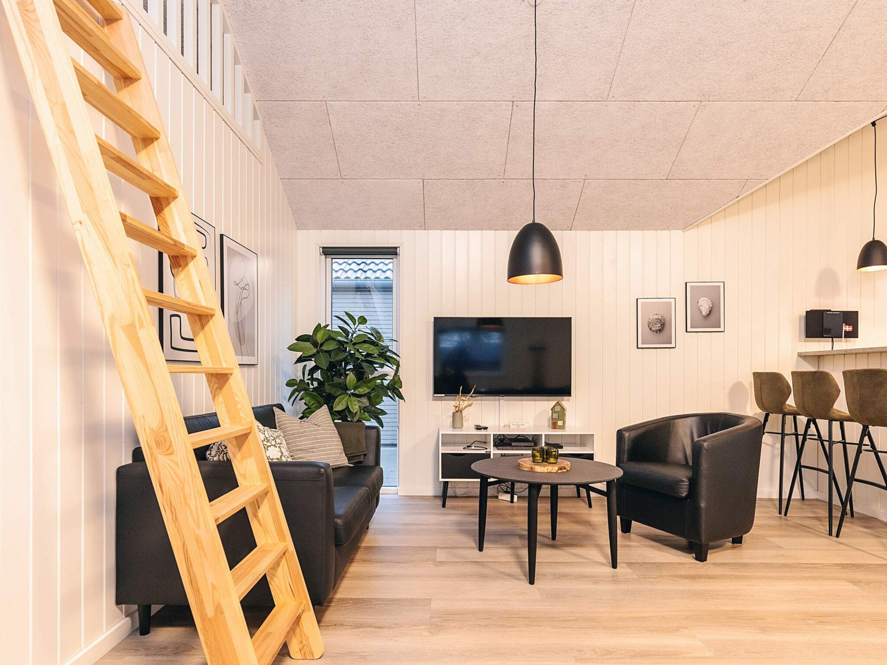 Un salon moderne avec une échelle en bois, un canapé noir, une table basse ronde, un fauteuil en cuir noir et un téléviseur fixé au mur.