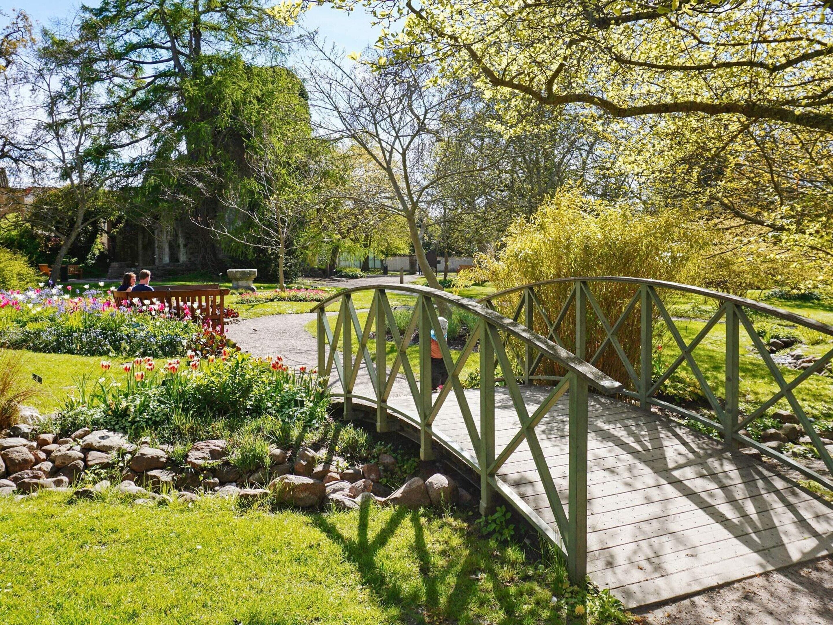 A wooden bridge over a garden with a variety of flowers and a stone wall in the background.