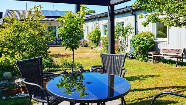 A patio with a round table and four chairs, surrounded by a garden with trees and a building in the background. - 4 Star Holiday Home in Visby (Visby)