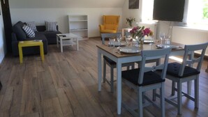 Een moderne woonkamer met een bank, een gele fauteuil, een eettafel gedekt voor twee personen en een flatscreen televisie aan de muur.