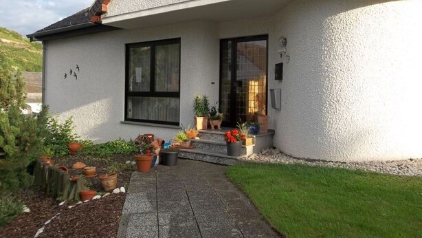 A house with a white exterior, a black-framed window, and a door with a glass panel. There are potted plants and a small garden area in front. - in Landhuisstijl met Uitzicht op de Moezel (Piesport)