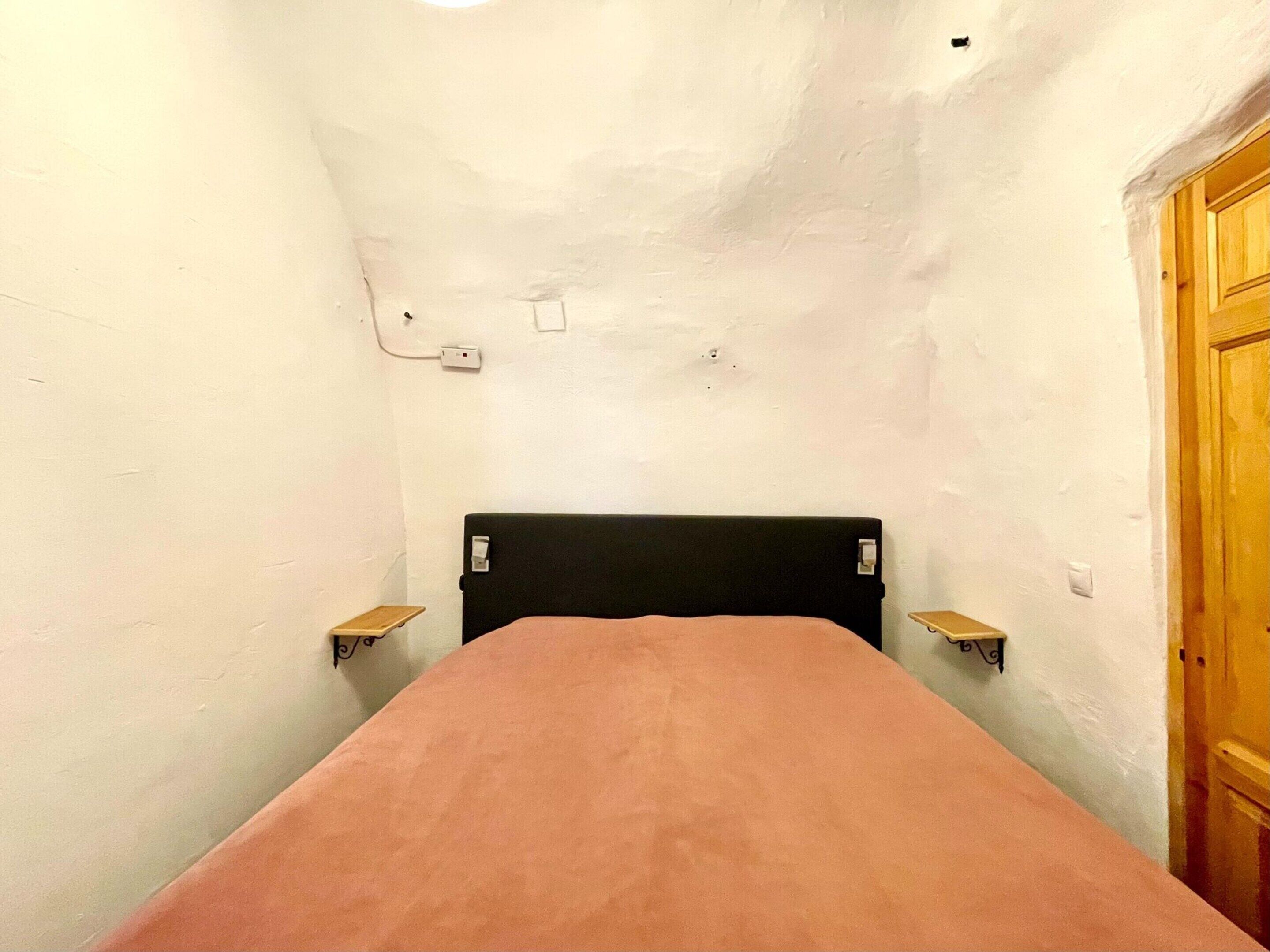 A narrow room with a single bed, a wooden door, and a small shelf.