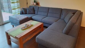 A modern living room with a grey sectional sofa, a wooden coffee table, and a green mat. There is a potted plant on a small shelf and a floor lamp. - Holiday Home Manders (Wangerland)