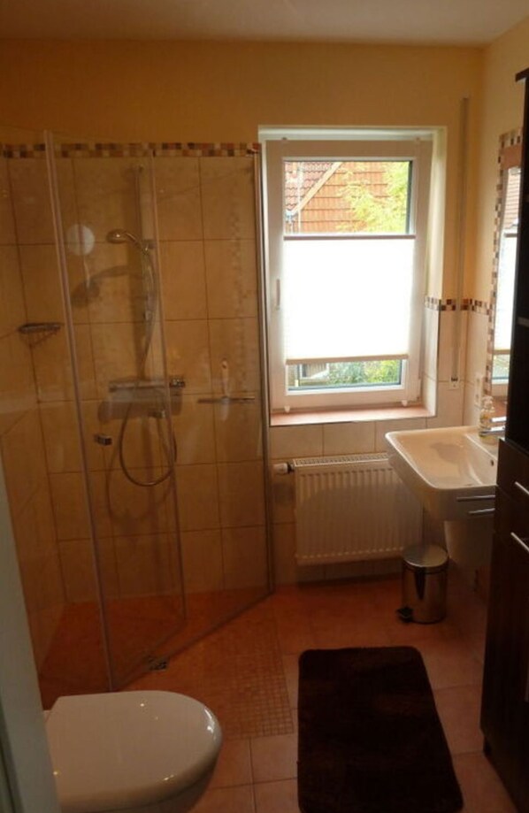 A bathroom with a glass shower enclosure, a toilet, a sink, and a window providing natural light. - Holiday Home Manders (Wangerland)