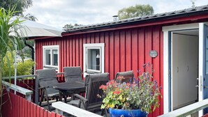 A red building with a white door and windows, a wooden deck with chairs and a table, and a large blue planter with flowers. - 4 Star Holiday Home in Solvesborg (Sölvesborg)