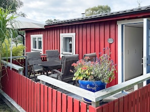 Ein rotes Gebäude mit einer weißen Tür und Fenstern, eine Holzterrasse mit Stühlen und einem Tisch und ein großer, blauer Pflanzkasten mit Blumen.