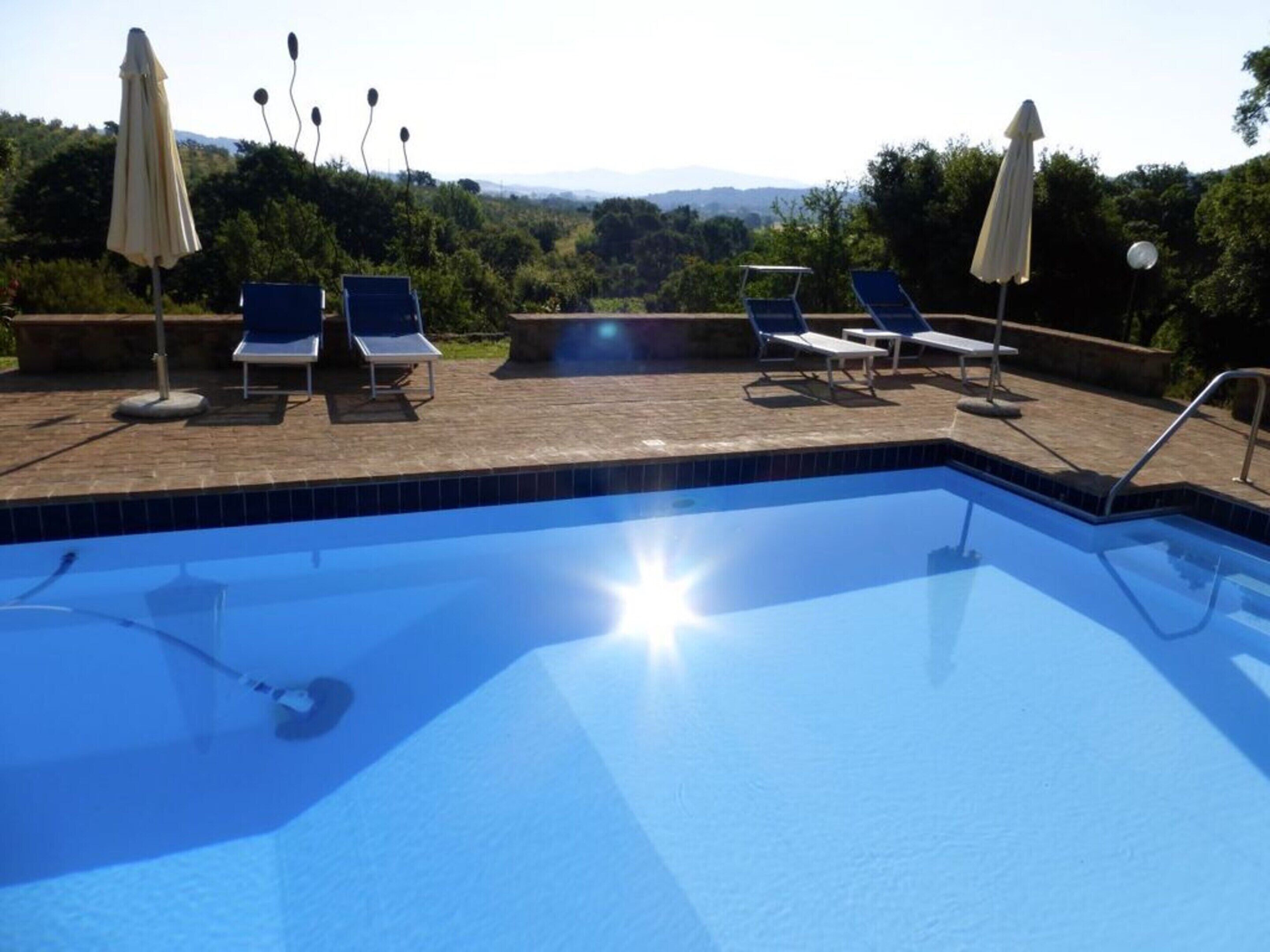 Una piscina con sillas de descanso y sombrillas, rodeada de vegetación y vistas a colinas.
