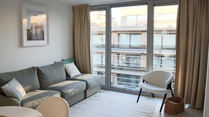 A modern living room with a sofa, a chair, and a large window with curtains. - Apartment in Nieuwpoort Near the Beach (Nieuwpoort)