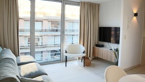 A modern living room with a sofa, a chair, a TV, and a large window with curtains. - Apartment in Nieuwpoort Near the Beach (Nieuwpoort)