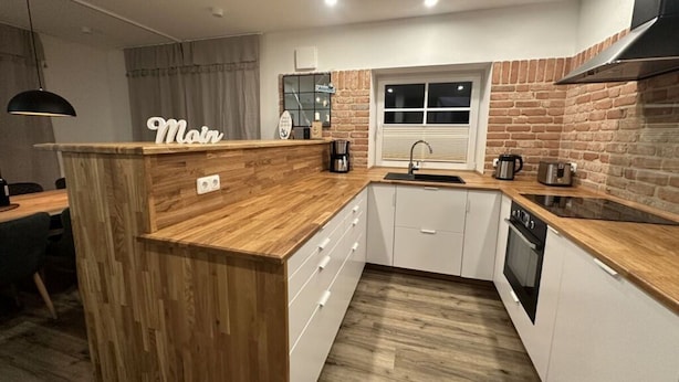 A modern kitchen with a wooden island, white cabinets, a brick accent wall, and a window above the sink. - Eiderhuus Modern Retreat (Toenning)