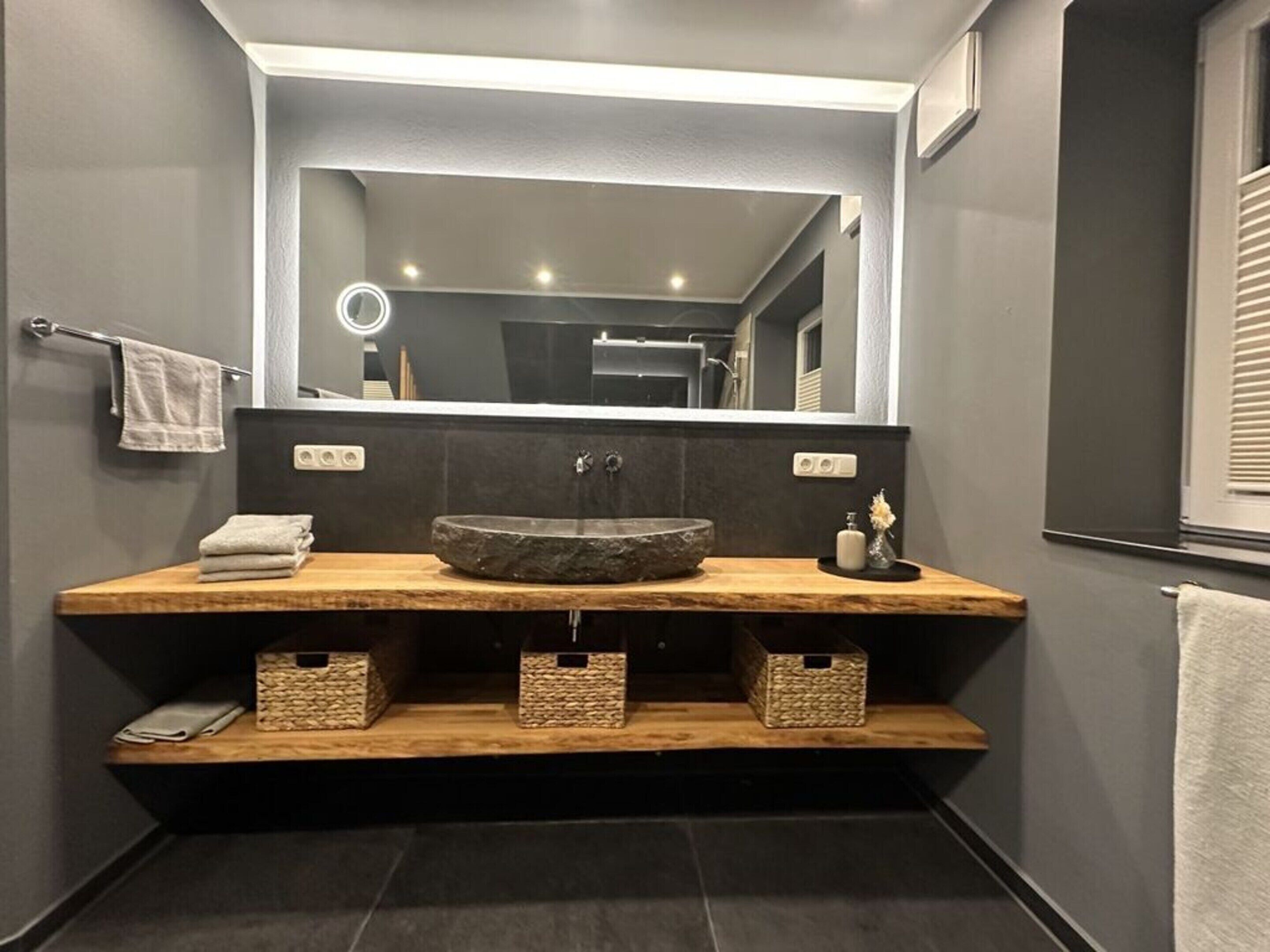 A modern bathroom with a large mirror, a central sink, and wooden storage shelves.