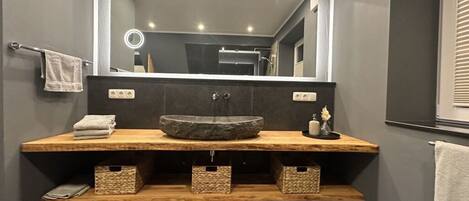 A modern bathroom with a large mirror, a central sink, and wooden storage shelves.
