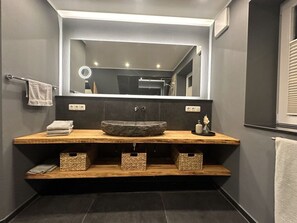 A modern bathroom with a large mirror, a central sink, and wooden storage shelves. - Eiderhuus Modern Retreat (Toenning)