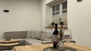 A modern living room with a wooden table, two wine glasses, and a bottle of Merlot. There is a gray sofa and a small round table with a decorative item. A window with blinds and a shelf with plants is visible in the background. - Eiderhuus Modern Retreat (Toenning)