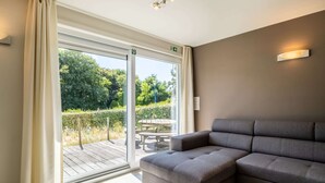 Ein modernes Zimmer mit einem grauen Sofa, einem Holztisch und einer Glastür, die zu einem Außenbereich mit einer Bank und Grünpflanzen führt.