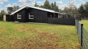 Un bâtiment noir avec un toit en pente, une cour clôturée et des arbres en arrière-plan.