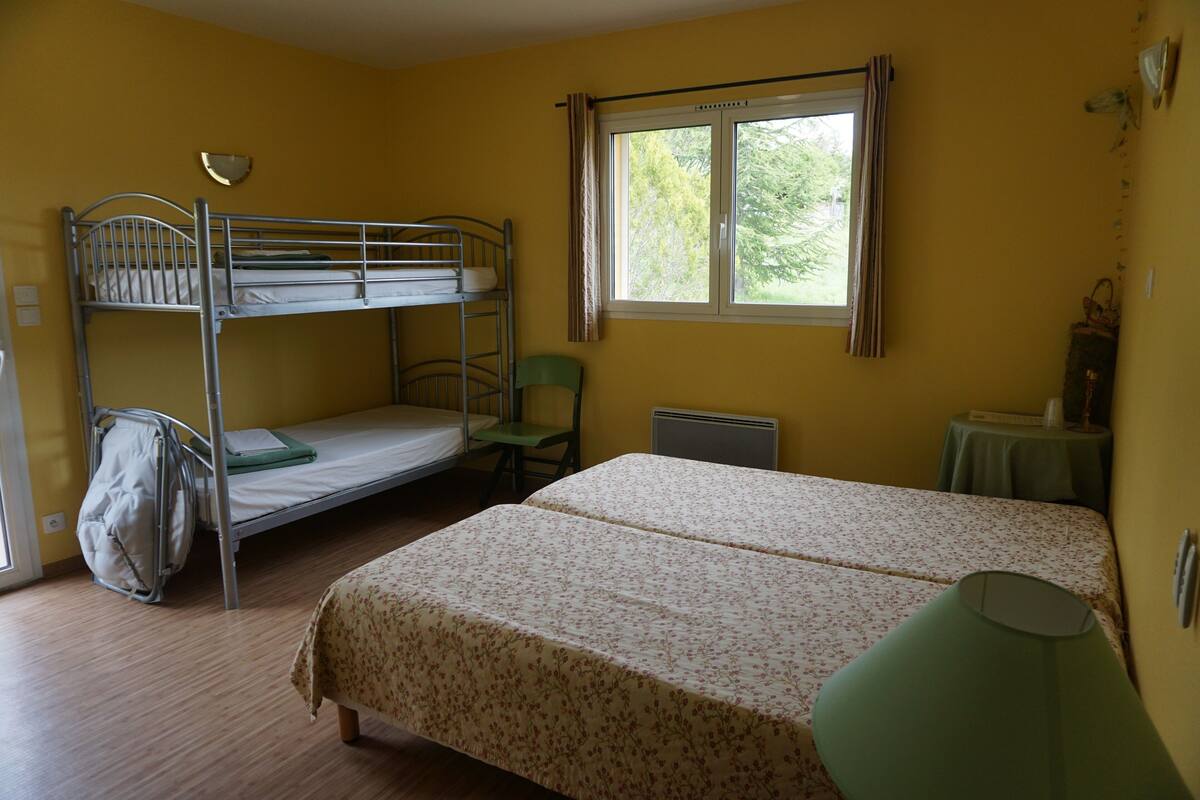 A room with a bunk bed, a single bed, a chair, a table, and a window with curtains.