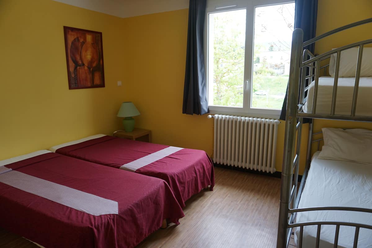 A room with two single beds, a bunk bed, a radiator, a window with curtains, a lamp, and a painting on the wall.